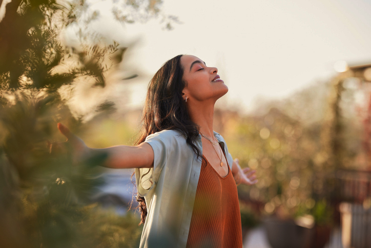 Young latin woman with arms outstretched breathing in fresh air during sunrise at the balcony. Healthy girl enjoying nature while meditating during morning with open arms and closed eyes. Mindful woman enjoying morning ritual while relaxing in outdoor park.